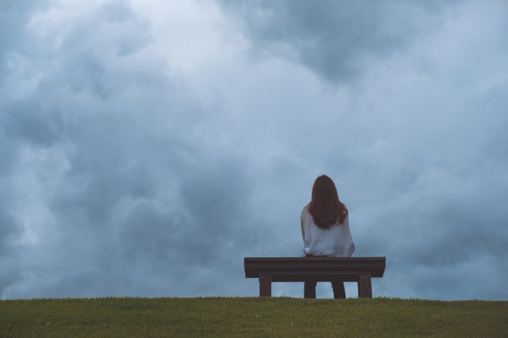 A woman sitting alone on a wooden bench in the park with cloudy
