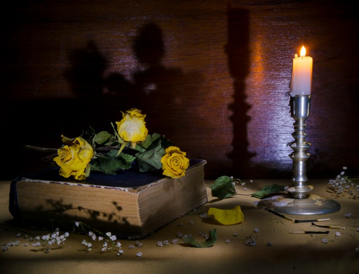 Still life old yellow roses lying on the book, a candlestick with a burning candle.
