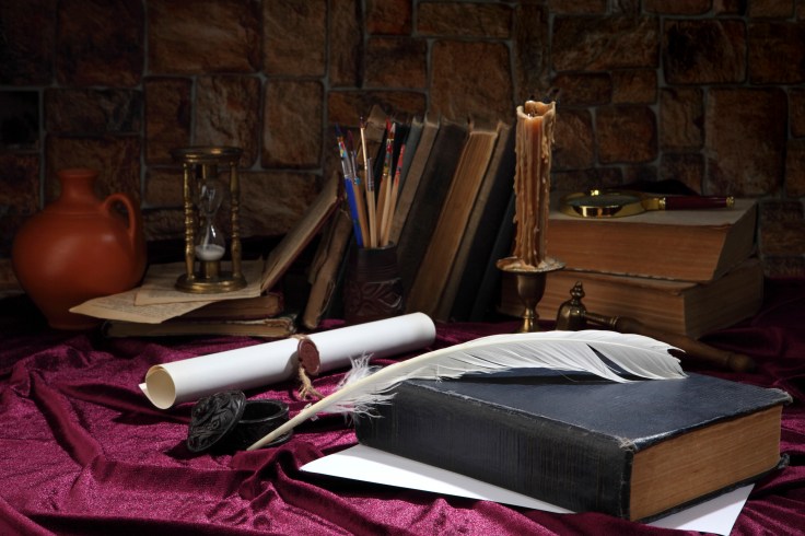A goose feather, an inkwell, a scroll with a seal, a forged bronze candlestick with a candle, books, a magnifying glass and an hourglass on the desktop. Retro stylized photo. Selective focus.